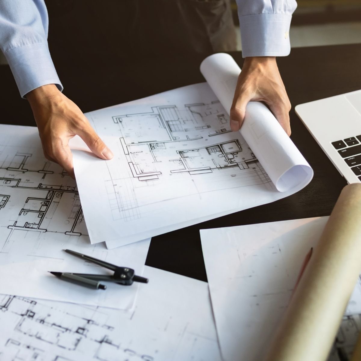 Civil engineer unrolling and reviewing detailed architectural floor plan blueprints on a desk with drafting compass and laptop representing ATP Development & Construction's comprehensive civil engineering services
